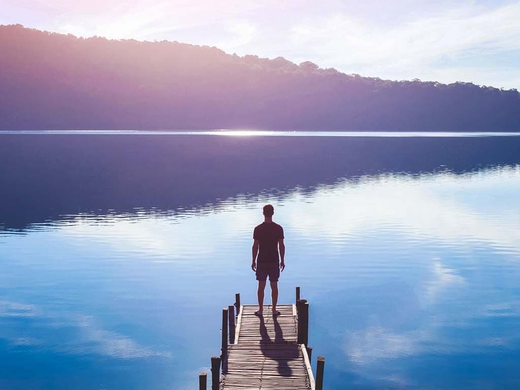 Henkilö seisoo laiturilla katsoen kohti peilityyntä järveä ja vuoristoista horisonttia.
