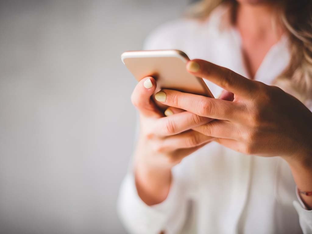 Nainen pitää älypuhelinta molemmin käsin ja käyttää sitä, pukeutuneena vaaleaan paitaan, taustalla pehmeästi sumennettu seinä.