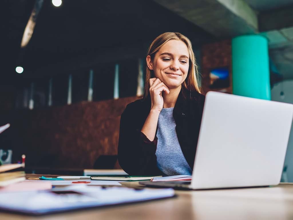 Nainen istuu nykyaikaisessa toimistoympäristössä kannettavan tietokoneen ääressä, hymyilee ja nojaa käteensä työpöydän äärellä.