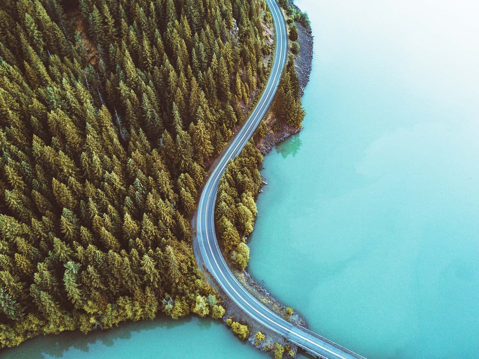 Ilmakuva mutkittelevasta tiestä, joka kulkee tiheän metsän halki turkoosin järven rannalla kirkkaassa säässä.