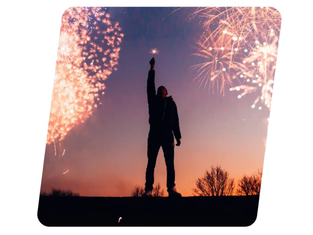 Henkilö seisoo iltahämärässä käsi voitokkaasti ilmaan ojennettuna, taustalla ilotulituksia värikkäällä taivaalla.