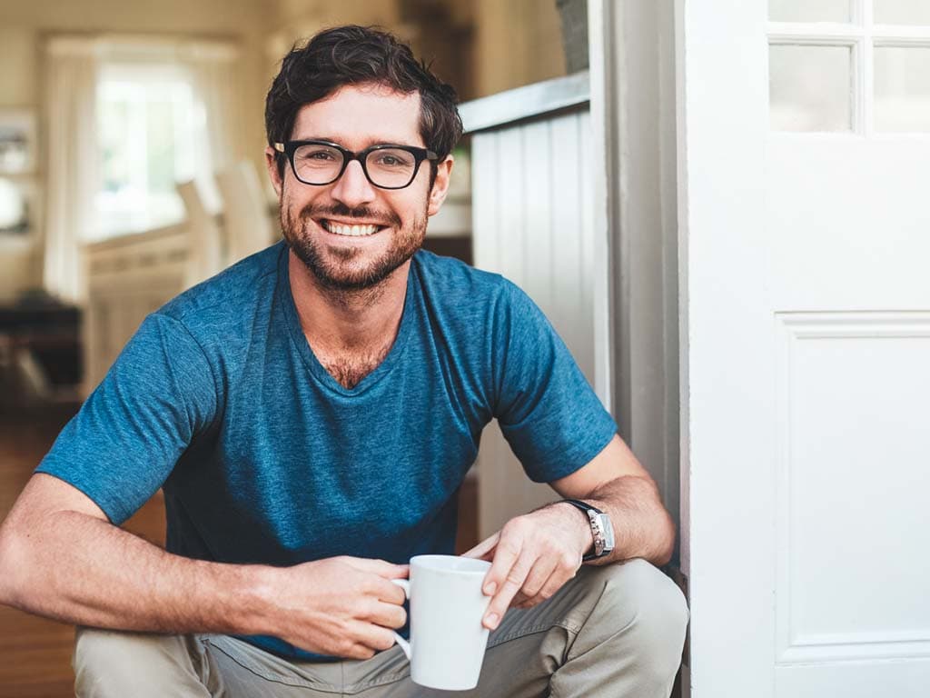 Mies istuu oven vieressä rentona ja hymyilee, kädessään valkoinen kahvikuppi kodikkaassa ympäristössä.