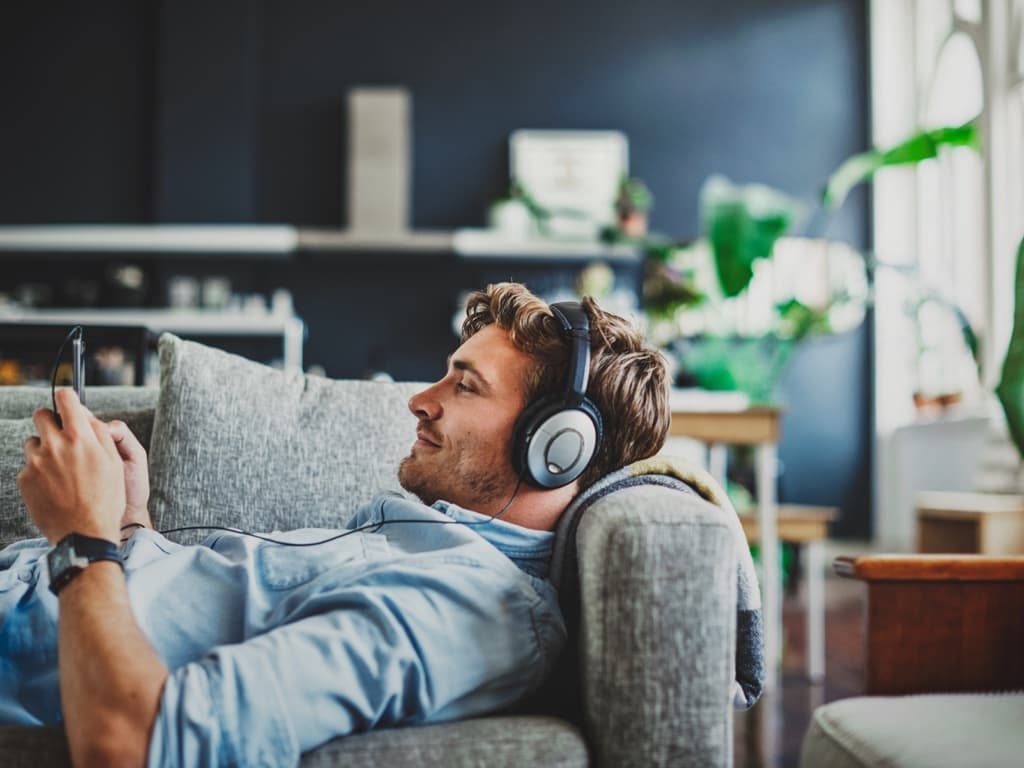 Mies makaa sohvalla kuulokkeet korvillaan ja käyttää älypuhelinta modernissa olohuoneessa.